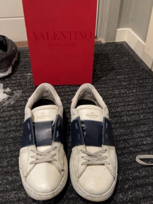 Valentino Garavani vita och marinblå sneakers - Säljer ett par snygga Valentino Garavani sneakers i vitt och marinblått skinn. Skorna har klassisk låg profil, rund tå och bred marinblå detalj över mitten. De har vita snören och diskret Valentino-logga på plösen. Kommer med originalkartong.