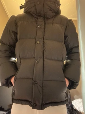 Svart pufferjacka med huva - Säljer en svart pufferjacka med huva och knappar framtill. Jackan har dragkedjor på fickorna och snörning vid huvan. Insidan har ett rutigt foder i huvan som ger en snygg kontrast. Perfekt för kalla dagar och riktigt skön att bära.