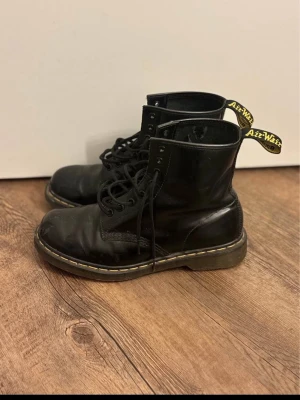 Svarta Dr. Martens boots - Klassiska svarta Dr. Martens boots i skinn med snörning och den ikoniska gula sömmen runt sulan. Grov sula och dragflik baktill med gul text. Perfekta för dig som vill ha en tidlös och edgy stil.