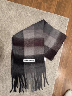 Rutig Acne Studios halsduk i ull - Säljer en stor och fluffig halsduk från Acne Studios i rutigt mönster med mörkgrå, svart och vinröda toner. Halsduken har långa fransar i ändarna och en tydlig Acne Studios-logga. Perfekt accessoar för kalla dagar.