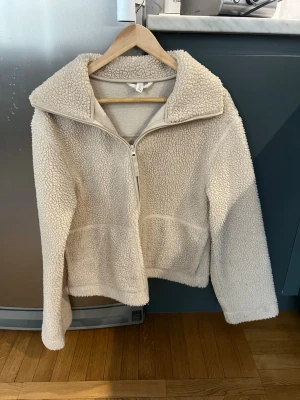 Teddyjacka med dragkedja - Mysig beige teddyjacka från H&M med hög krage och hel dragkedja framtill. Jackan är fluffig och har en avslappnad passform, perfekt för kyliga dagar. Materialet är mjukt, den är väl använd men i väldigt bra skick
