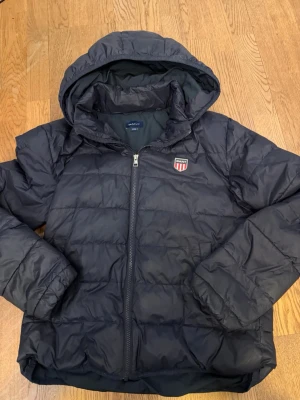 Gant jacka  - Säljer en mörkblå pufferjacka från Gant med huva och dragkedja framtill. Jackan har ett broderat Gant-märke på bröstet och praktiska fickor med tryckknapp. Perfekt för kalla dagar och riktigt skön att bära.