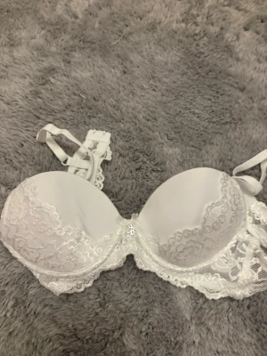 Vit spets-bh U Bella, A70 - Säljer en vit bh från U Bella i storlek A70. Bh:n har vadderade kupor och är dekorerad med vacker spets och en liten rosett framtill. Axelbanden är justerbara och knäppningen bak har flera hakar för bästa passform.