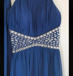 Marinblå långklänning med paljettdetaljer - Säljer en marinblå långklänning med glittriga paljettdetaljer i midjan och runt halsen. Klänningen har ett elegant fall, är ärmlös och har en hög ringning. Perfekt för bal eller festliga tillfällen. Materialet känns lätt och flytande.