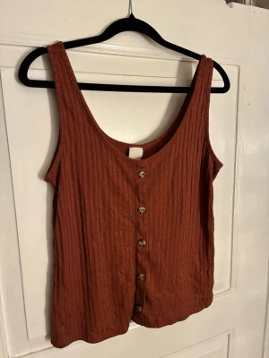 Rostbrun ribbad linnetopp från H&M - Säljer en rostbrun ribbad linnetopp från H&M i storlek S. Toppen har knappar framtill och är ärmlös med breda axelband. Materialet är mjukt och stretchigt, perfekt för varma dagar. Enkel och snygg att matcha med jeans eller kjol.