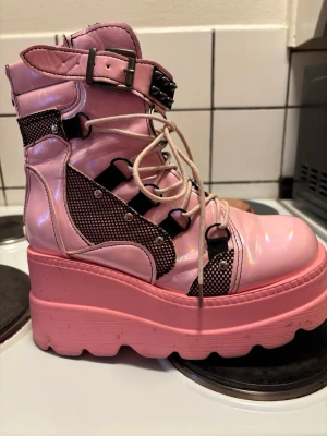 Rosa platåboots med snörning - Unika rosa boots med hög platåsula och snörning. Skorna har svarta meshdetaljer, metallnitar och ett spänne med nitar upptill. Tillverkade i glansigt syntetmaterial och har en chunky, edgy look som sticker ut.
