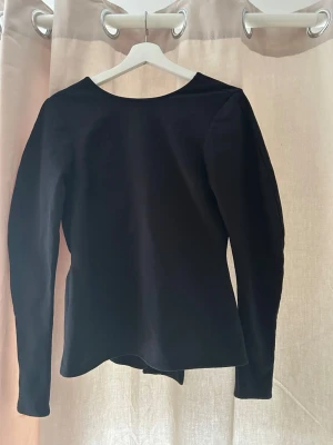 Svart långärmad omlottopp med knyt - Snygg svart långärmad topp med omlottdesign och knyt framtill. Toppen har en djup v-ringning och är figurnära i modellen. Perfekt att styla med jeans eller kjol för en trendig look.