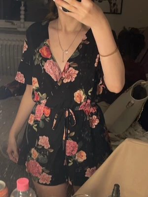 Svart blommig byxdress med knyt - Säljer en svart byxdress med färgglatt blommönster i rosa, rött och grönt. Byxdressen har korta ärmar, v-ringning och ett knytband i midjan för en snygg siluett. Perfekt för dig som vill ha något bekvämt men ändå stilrent. Står inte storlek, men gissar på S