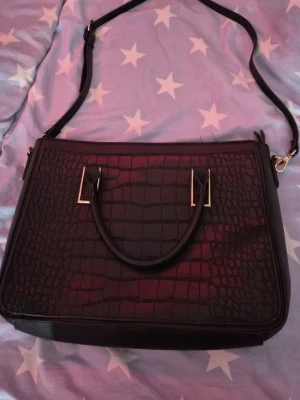 Svart axelväska från Anna Field - Snygg svart axelväska från Anna Field med krokopräglat mönster på framsidan och slät baksida. Väskan har både handtag och justerbar axelrem samt guldfärgade detaljer. Invändigt finns dragkedjefack för småsaker. Perfekt för dig som gillar stilrena accessoarer.