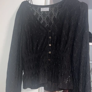 Tröja  - Säljer en svart blus från Holly & White med v-ringning och knappar framtill. Blusen har ett diskret rutmönster och är tillverkad i ett lätt, transparent tyg. Långärmad modell med smockad midja som ger en snygg siluett. Tröjan har bara använts ett fåtal gånger och funkar även för dig som har storlek S 