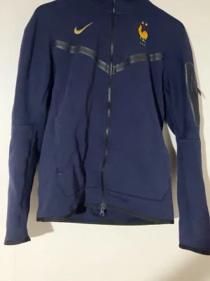 Mörkblå Nike hoodie FFF herr S - Säljer en mörkblå hoodie från Nike med FFF-logga och broderad tupp på bröstet. Tröjan har hel dragkedja, lång ärm och guldfärgade detaljer. Tillverkad i mjukt material och har en sportig look med svarta band över bröstet.