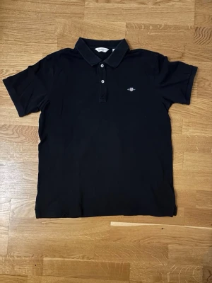 Svart piké från GANT, 176 cm - Klassisk svart pikétröja från GANT med korta ärmar och liten logga broderad på bröstet. Tröjan har krage och knappar framtill för en clean look. Perfekt till jeans eller shorts för en enkel men snygg stil. OBS 170/176 köpt som en 176 men känns som 170