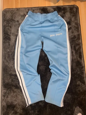 Palm Angels ljusblå trackpants - Säljer ett par ljusblå trackpants från Palm Angels med vita och svarta ränder längs sidorna. Byxorna har elastisk midja, raka ben med dragkedja vid benslut och Palm Angels-logga tryckt på vänster sida. Tillverkade i 100% polyester.