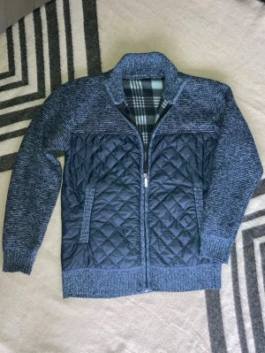 Cardigan från Fashton BB - Säljer en blå puffer med quiltad framsida och stickade ärmar samt rygg. Jackan har dragkedja framtill, ribbade muddar och krage. Insidan har ett rutigt foder i svart, vitt och grått. Perfekt för dig som vill ha en unik och varm jacka.
