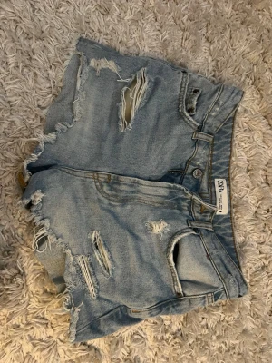 Ljusa denimshorts från Zara - Säljer ett par ljusblå denimshorts från Zara med hög midja och råa, slitna detaljer. Shortsen har flera revor och fransiga kanter för en riktigt avslappnad vibe. Klassisk femficksmodell och knappgylf. Perfekta till sommaren!