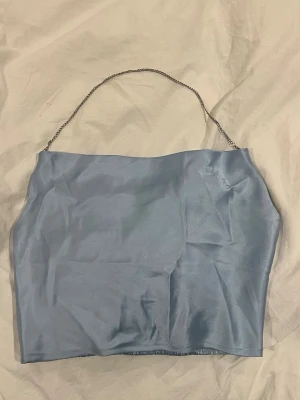 Ljusblå satin bandeau topp med kedjeband - Snygg ljusblå bandeau topp i satin med smalt kedjeband som axelband. Toppen är axelbandslös och har smockad baksida för extra komfort och passform. 