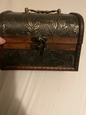 Mörkbrun smyckeskrin med blommönster - Unik handväska i mörkbrunt blommönster och snygga trädetaljer. Väskan har ett dekorativt metallhandtag och klassiskt spänne framtill. Perfekt för dig som gillar vintage vibes och vill sticka ut med en cool accessoar.