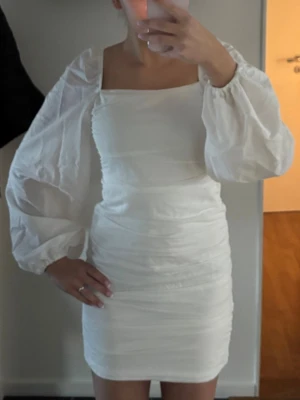 Vit kort klänning med puffärmar - Supersnygg vit kortkläning med puffiga långärmar och fyrkantig urringning från Nelly. Klänningen har en tight passform med rynkade detaljer längs hela kroppen och är tillverkad i ett tunt och lätt material. Använd 1 gång🤍