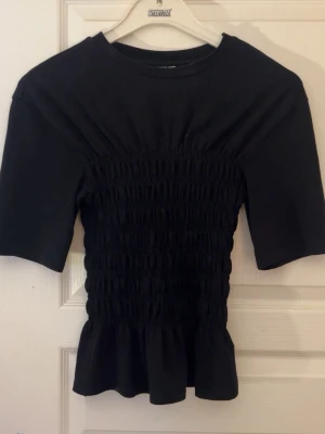 Svart smockad t-shirt & Other Stories - Supersnygg svart t-shirt från & Other Stories med smockad midja och volangkant nedtill. Modellen har korta ärmar och rund halsringning. Perfekt till jeans eller kjol för en trendig look.