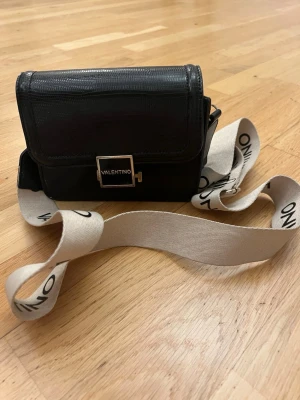 Svart axelväska från Valentino - Snygg svart axelväska från Valentino med strukturmönstrad yta och tre olika axelband – ett brett beige band med logga och ett smalare svart band och ett kedjeband. Väskan har ett stilrent metallspänne framtill och fack inuti för smart organisering.