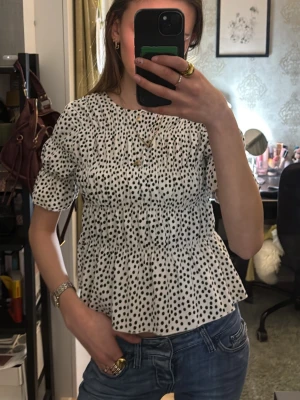 Vit prickig blus med puffärm - Supersöt vit blus med svarta prickar och puffiga korta ärmar. Blusen har en avslappnad passform med rynkade detaljer och volang nedtill🥰 (OBS. tajt vid kragen som gör det svårt att få på men ej tajt på❤️) Kids size men räknas som XS