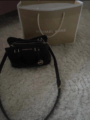 Svart handväska från Michael Kors - Säljer en snygg svart handväska från Michael Kors i äkta skinn. Väskan har guldfärgade detaljer, dubbla handtag och en avtagbar axelrem. Praktisk dragkedja upptill och klassisk MK-logga framtill. Perfekt storlek för det viktigaste och stilren design.