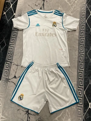 Real Madrid fotbollskit Ronaldo #7 - Säljer ett vitt Real Madrid matchställ från Adidas med blå detaljer. Tröjan har klubbmärke, Fly Emirates-tryck och tre blå ränder på axlarna. Baksidan har handskrivet 'Ronaldo' och nummer 7. Shortsen har blå ränder och klubbmärke. Perfekt för fotbollsälskare!