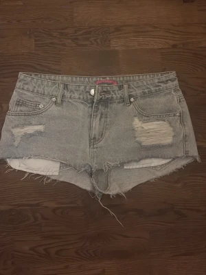 Blå denimshorts från Edikted - Säljer ett par gråa denimshorts från Edikted med råa kanter och slitna detaljer framtill. Klassisk femficksmodell med knapp och dragkedja. Perfekta för sommaren och ger en avslappnad vibe.