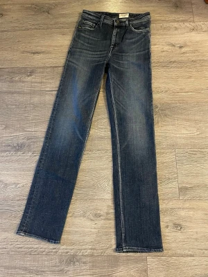 Mörkblå jeans från Tiger of Sweden - Säljer ett par mörkblå jeans från Tiger of Sweden med klassisk femficksdesign och raka ben. Jeansen har snygga slitningar och subtil tvätt för en cool look. Tillverkade i mjukt jeansmaterial med knappgylf och bälteshällor. 