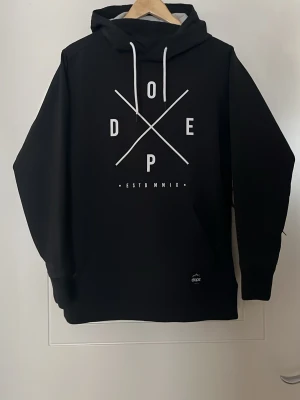 Svart skidjacka från Dope - Säljer en svart skidjacka/hoodie från Dope med stor vit logga och grafiskt tryck på bröstet. Jackan har huva med snörning, stor ficka med dragkedja på magen och dragkedjor under armarna för ventilation. Den har även fleece inuti. Använd några gånger men är i mycket gott skick! Priset är inte hugget i sten.