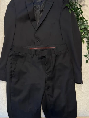 Svart barn kostym slim fit Dressmann - Säljer en svart kostym från Dressmann i slim fit-modell.  Kavajen har klassisk krage, enkel knappstängning och matchande byxor med bälteshällor.   Kavajens storlek : 42-short  Byxornas storlek: W28/L30  Kostymen är i bra skick förutom att den skulle behöva rollas 😊😇  Tveka inte att höra av dig vid frågor!😁