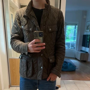 Quiltad höstjacka från Barbour M - Säljer en grön quiltad jacka från Barbour i storlek M. Jackan har snyggt rutmönster, flera fickor med tryckknappar och dragkedja framtill. Insidan har ett coolt tryck med motorcykelmotiv. Perfekt för dig som gillar klassisk stil med edge.