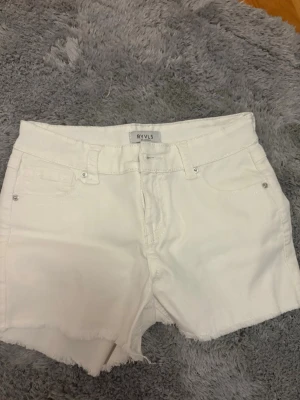 Vita jeansshorts från RYVLS - Snygga vita jeansshorts från RYVLS med råa kanter och klassisk femficksdesign. Shortsen har hög midja, bälteshällor och silverfärgade nitar. Perfekta för varma dagar och enkel att matcha med allt.