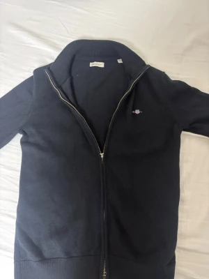 Svart kofta från GANT med dragkedja - Klassisk svart kofta från GANT med hel dragkedja framtill och diskret logga på bröstet. Tröjan har ribbad krage och muddar, samt är långärmad. Perfekt till lager på lager och enkel att matcha med allt.