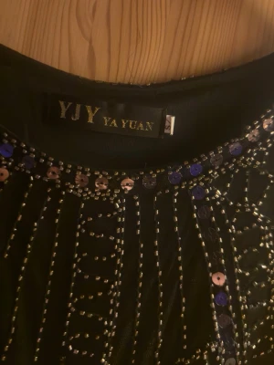 Svart paljettklänning från YA YUAN - Supersnygg svart kortklänning från YA YUAN med glittriga paljetter och pärldetaljer i geometriskt mönster. Klänningen är ärmlös och har en kroppsnära passform som verkligen sticker ut. Perfekt för dig som vill glänsa extra mycket!