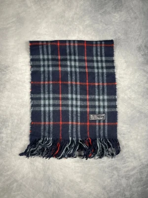 Rutig ull scarf från Burberry - Klassisk rutig scarf från Burberrys i marinblått, grått och rött. Tillverkad i 100% lammull med fransar nedtill och tydlig Burberrys-label. Perfekt för dig som gillar tidlös brittisk stil och vill ha något extra till din outfit. 