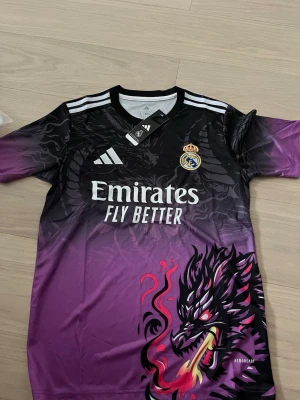 Real Madrid x Adidas draktröja S - Säljer en limited edition Real Madrid x Adidas tröja i storlek S med coolt drakmotiv. Tröjan har svart och lila toning, vita Adidas-ränder på axlarna och Real Madrid-logga på bröstet. Materialet är lätt polyester och passformen är slim fit. Perfekt för dig som gillar unika fotbollströjor.