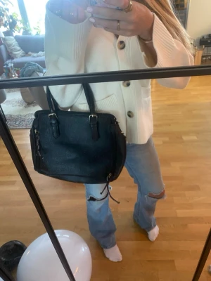 Svart handväska i skinnlook - Säljer en stilren svart handväska med två rejäla handtag och detaljer i silverfärgad metall. Väskan har en fyrkantig form och dragkedja upptill samt extra fack på sidorna. Perfekt storlek för vardagens essentials och enkel att matcha till olika outfits.