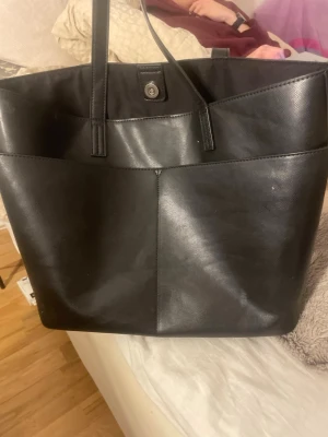 Svart shoppingväska i skinnimitation - Stor svart shoppingväska från Don Donna i skinnimitation med två rejäla axelband och magnetknäppning. Insidan har en praktisk ficka med dragkedja för småsaker. Perfekt när du vill ha med dig mycket grejer och ändå se snygg ut.