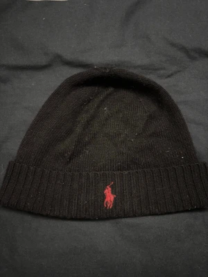 Svart mössa från Polo Ralph Lauren - Klassisk svart mössa från Polo Ralph Lauren i mjuk merinoull. Mössan har ribbad kant och är dekorerad med den ikoniska röda Polo-loggan broderad framtill. Perfekt accessoar för kyliga dagar.