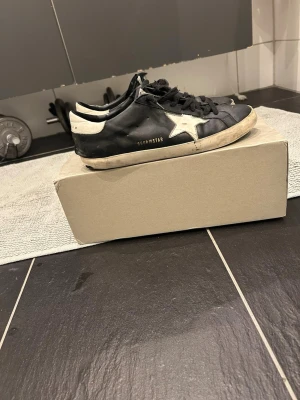 Golden Goose Superstar sneakers svart/vit - Golden Goose Superstar sneakers i svart skinn med vit stjärndetalj på sidan och vit häl. Klassisk låg modell med svarta skosnören och slitna detaljer för en cool, vintage look. Platt sula i gummi och rund tå. Perfekta för dig som gillar streetstyle. Defekter vid hörn märks inte vid användning 