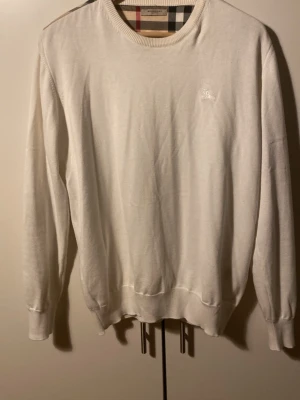 Vit Burberry tröja med rutiga detaljer - Säljer en vit tröja från Burberry med klassiska rutiga detaljer på axlar och armbågar. Tröjan har rund halsringning, långa ärmar och är tillverkad i mjuk bomull. Perfekt för dig som gillar stilrena plagg med en twist av lyx.