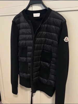 Svart quiltad jacka från Moncler - Säljer en svart quiltad jacka från Moncler med stickade ärmar och kropp i puffer-stil. Jackan har dragkedja framtill, två fickor och Moncler-logga på ärmen. Materialmix av polyester och stickat ger en snygg kontrast. Perfekt för dig som vill ha en stilren och modern look.