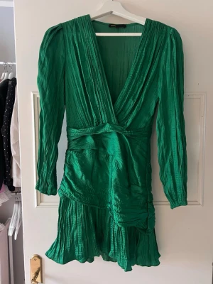 Grön volangklänning från Maje - Supersnygg grön kortklänning från Maje med   v-ringning. Klänningen har långa ärmar, volangdetaljer nertill och ett glansigt, plisserat tyg som ger en lyxig känsla. Perfekt för dig som vill sticka ut med färg och coola detaljer.