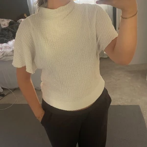 Blus  - Säljer en vit topp med ribbad struktur och korta ärmar. Toppen har en högre halsringning och sitter tajtare i midjan och runt magen. Den är stretchig i materialet.