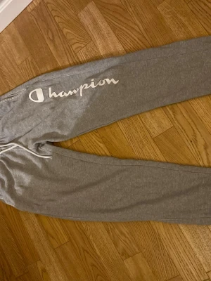 Grå mjukisbyxor från Champion - Säljer ett par grå mjukisbyxor från Champion med stort vitt logotryck längs ena benet. Byxorna har dragsko i midjan och muddar vid bensluten. Perfekta för chill eller träning, riktigt sköna och stilrena.
