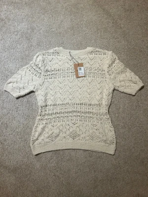 Stickad cremevit topp från ZARA - Säljer en cremevit stickad topp från ZARA med korta ärmar och ett snyggt hålmönster över hela plagget. Rund halsringning och ribbade kanter. Perfekt för lager på lager och ger en trendig touch till din outfit.