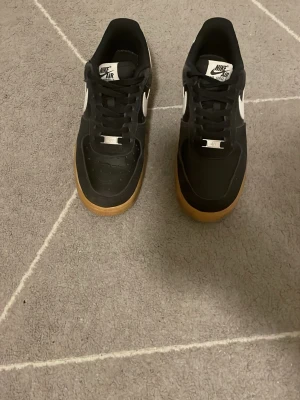 Svarta Nike Air Force 1 sneakers - Snygga svarta Nike Air Force 1 sneakers med vit swoosh och klassisk gummisula. Skorna har snörning och en låg siluett. Ovandelen är i syntetmaterial och skinn, vilket ger en stilren look. Perfekta för dig som gillar streetwear och tidlös design.