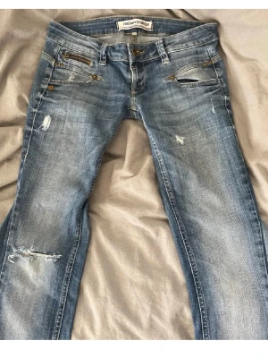 Låga bootcut jeans - Säljer ett par ljusblå bootcut jeans med slitna detaljer och låg midja. Säljer då de är för korta för mig.