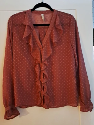 Rosa prickig blus med volanger - Söt rosa blus med små vita prickar och volangdetaljer längs den djupa V-ringningen och framtill. Blusen är tillverkad i ett tunt, transparent tyg och har långa ärmar med volang vid ärmslut. Perfekt för dig som gillar romantisk och trendig stil.